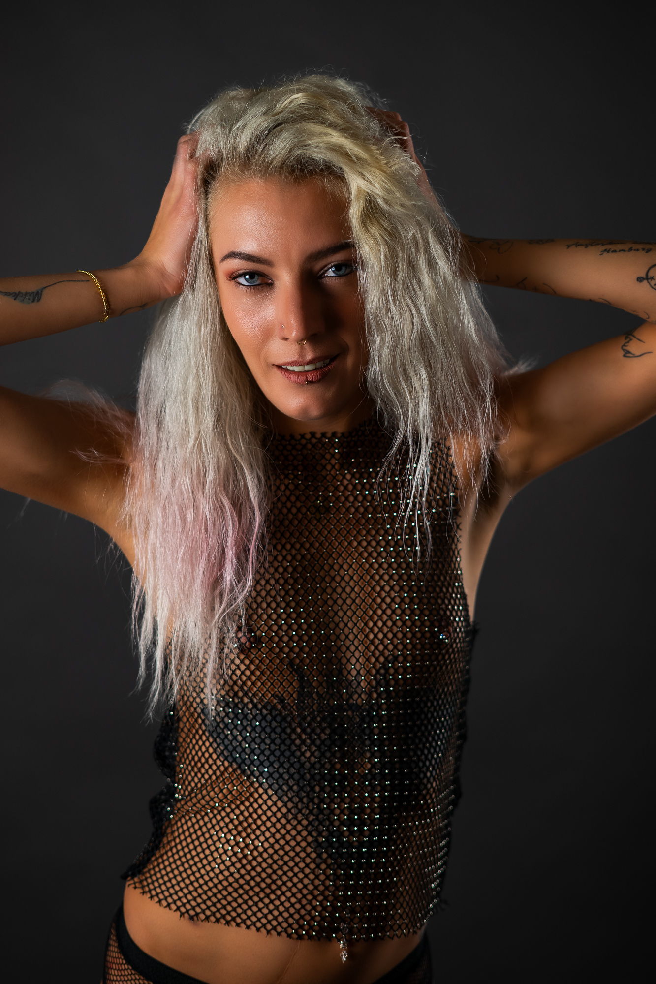 Tattoo-Model in goldgoldenem Mesh-Crop-Top, schwarzer Unterwäsche und Netzstrumpfhosen mit Overknee-Boots vor dunkelgrauem Studiohintergrund – Boudoir Portrait Fotografie
