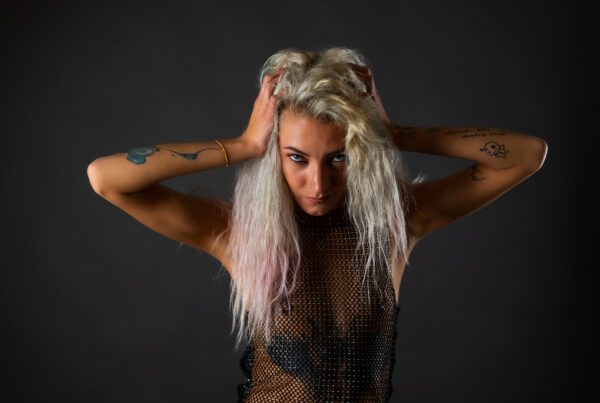 Tattoo-Model in goldgoldenem Mesh-Crop-Top, schwarzer Unterwäsche und Netzstrumpfhosen mit Overknee-Boots vor dunkelgrauem Studiohintergrund – Boudoir Portrait Fotografie