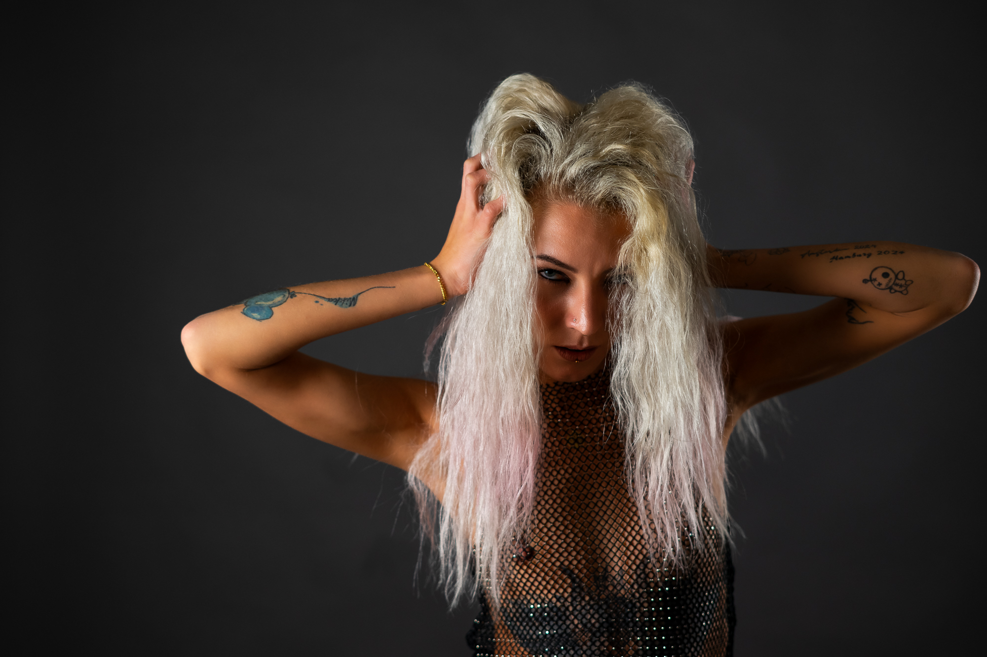 Tattoo-Model in goldgoldenem Mesh-Crop-Top, schwarzer Unterwäsche und Netzstrumpfhosen mit Overknee-Boots vor dunkelgrauem Studiohintergrund – Boudoir Portrait Fotografie