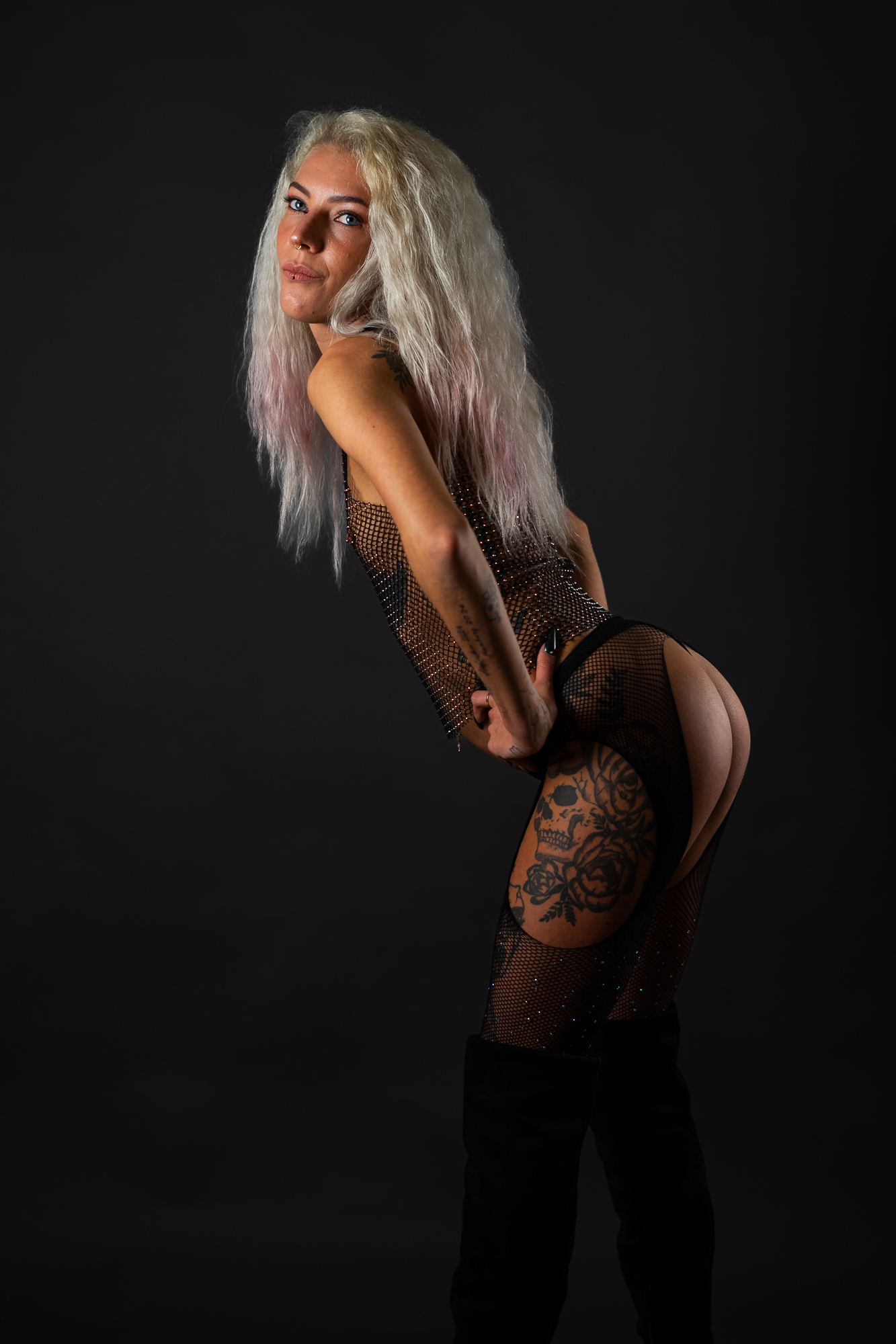 Tattoo-Model in goldgoldenem Mesh-Crop-Top, schwarzer Unterwäsche und Netzstrumpfhosen mit Overknee-Boots vor dunkelgrauem Studiohintergrund – Boudoir Portrait Fotografie